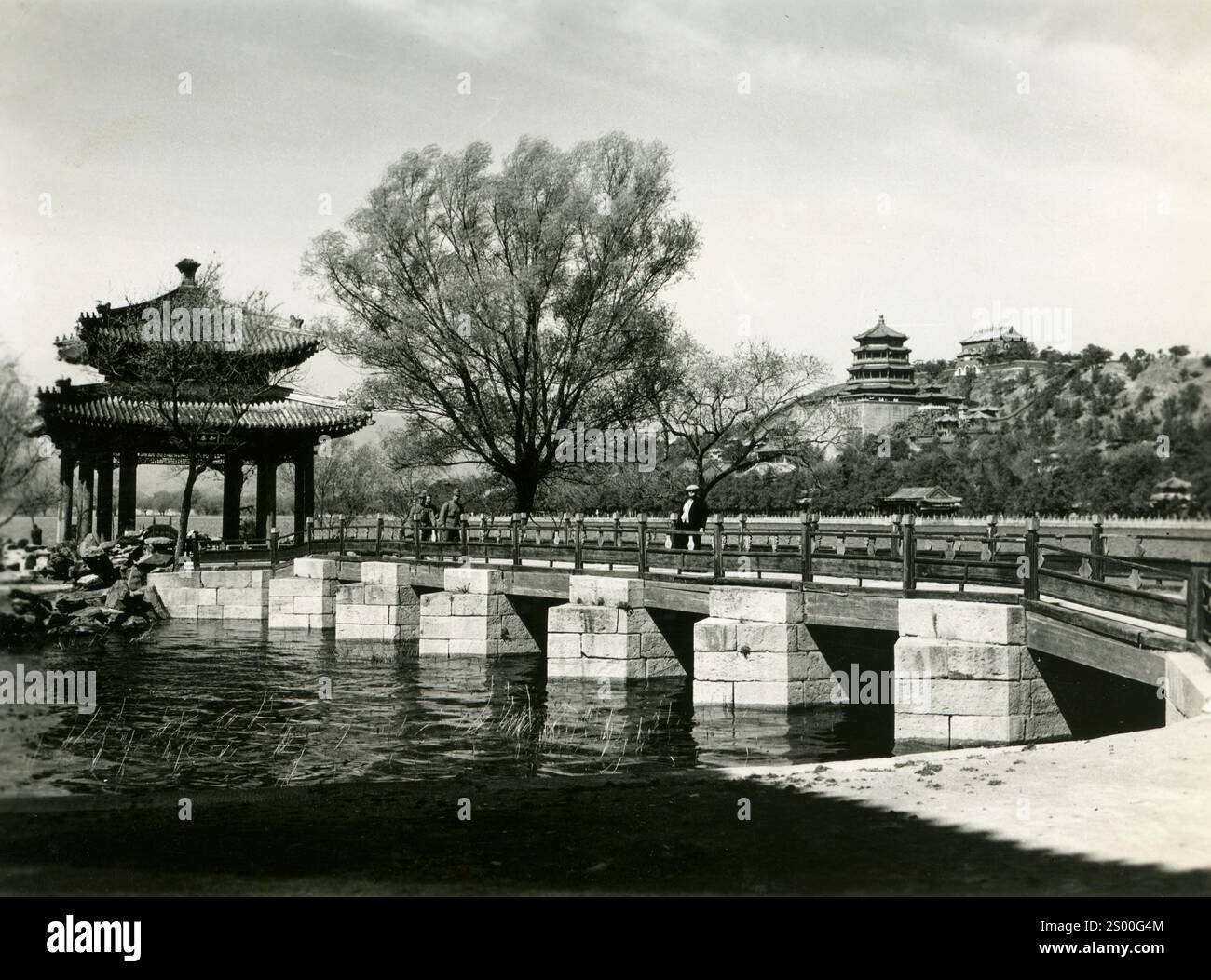 La bellezza senza tempo del Palazzo d'Estate: Ponte panoramico che attraversa acque tranquille con pagoda tradizionale e alberi ornamentali in uno storico paesaggio cinese. Pechino, Cina, anni 1930 Foto Stock