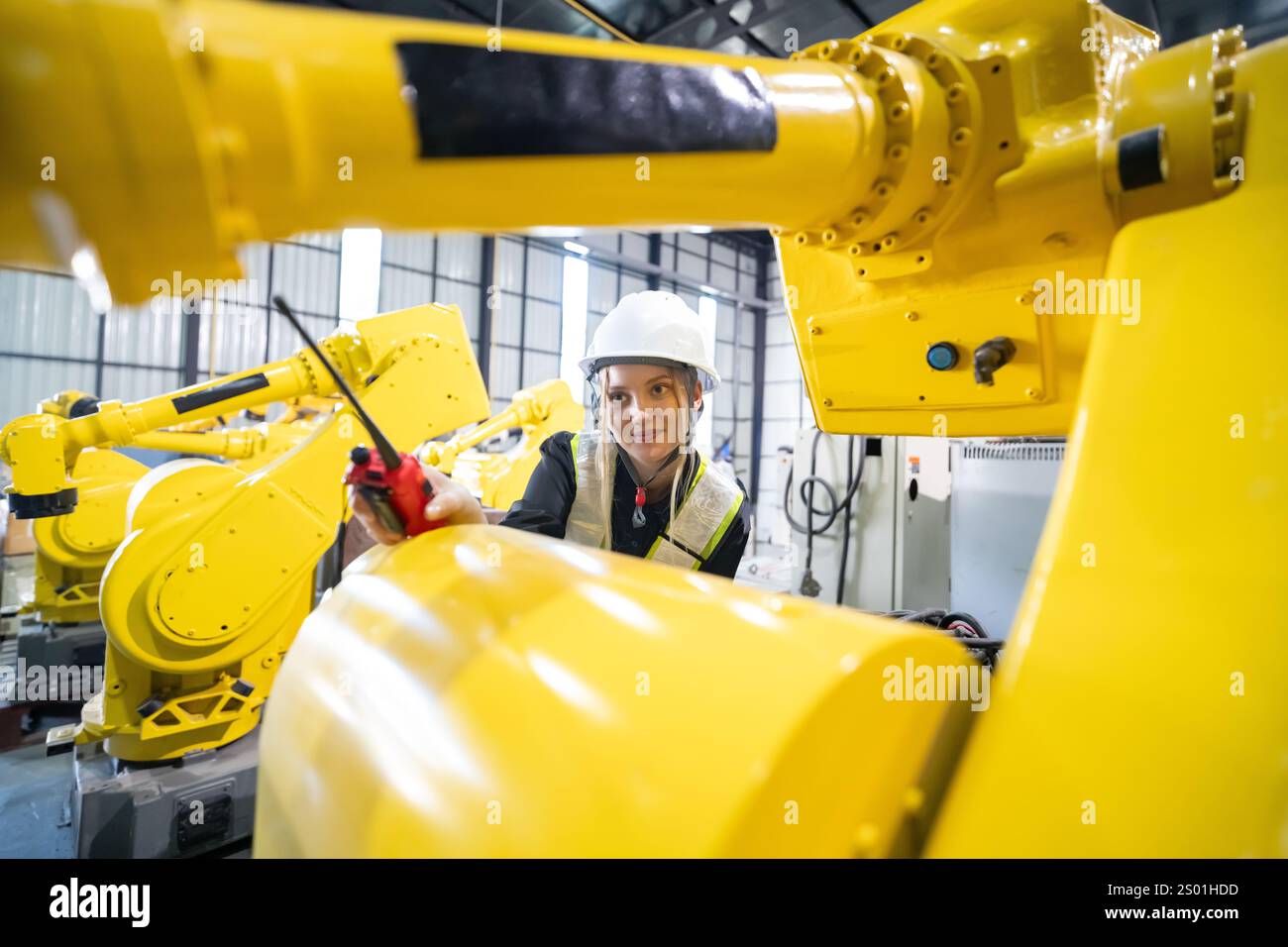 Un tecnico esperto, che indossa un elmetto e un equipaggiamento di sicurezza, lavora su bracci robotici gialli in un impianto di produzione molto affollato. L'ambiente mette in mostra l'avvento Foto Stock