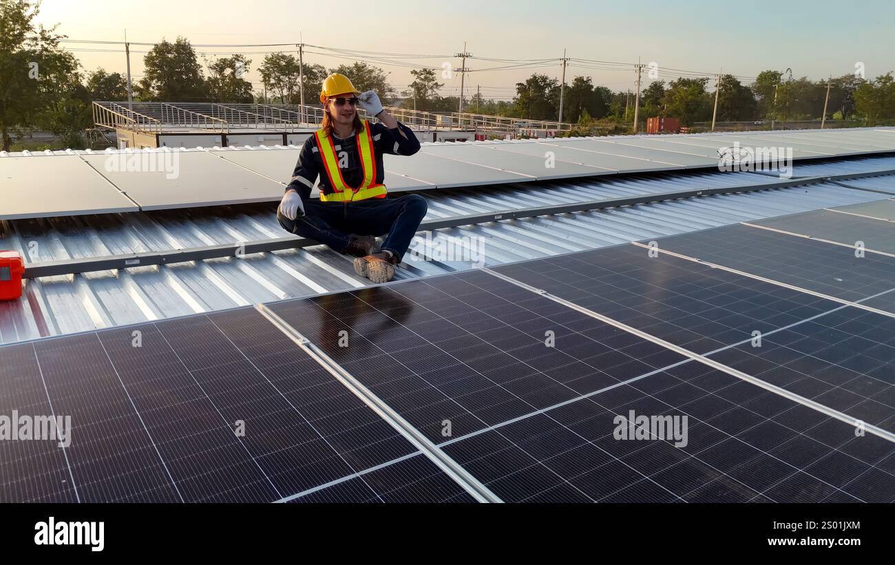 Un lavoratore con attrezzatura di sicurezza siede sul tetto di un pannello solare mentre parla al telefono. Foto Stock