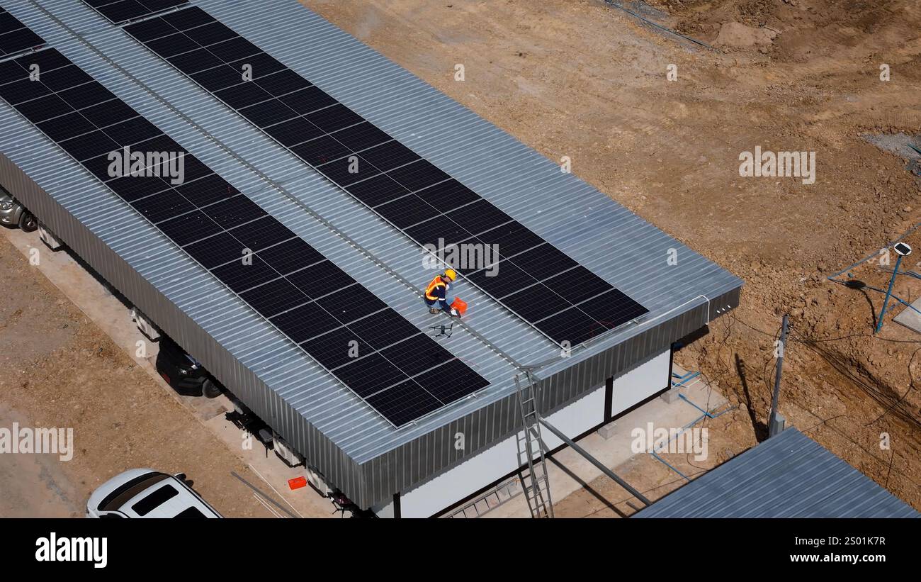 Due operai stanno installando pannelli solari sul tetto di un edificio commerciale sotto il cielo limpido. Il cantiere è caratterizzato da un paesaggio sabbioso e Foto Stock