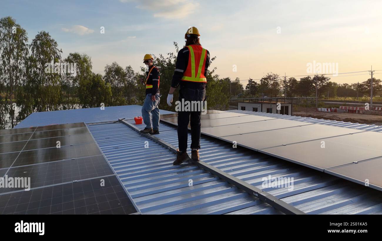 Due lavoratori stanno installando pannelli solari su un tetto durante il tramonto. Indossano caschi e giubbotti di sicurezza, dimostrando un impegno verso le energie rinnovabili Foto Stock