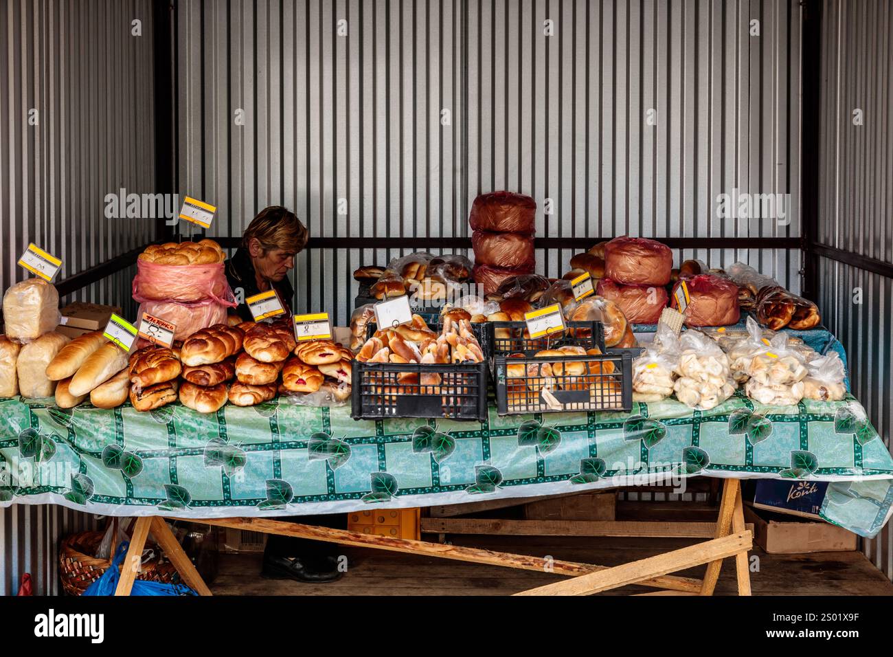 Una donna siede dietro un tavolo pieno di pane e pasticcini. Il tavolo è coperto da un panno blu ed è circondato da casse di prodotti da forno Foto Stock