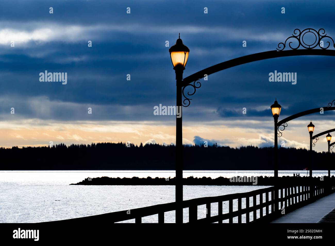 Le lampade sul molo di White Rock riflettono una banda di cieli assolati arancioni pallidi che appaiono a sud sotto bande di spesse nuvole tempestose blu e grigie Foto Stock