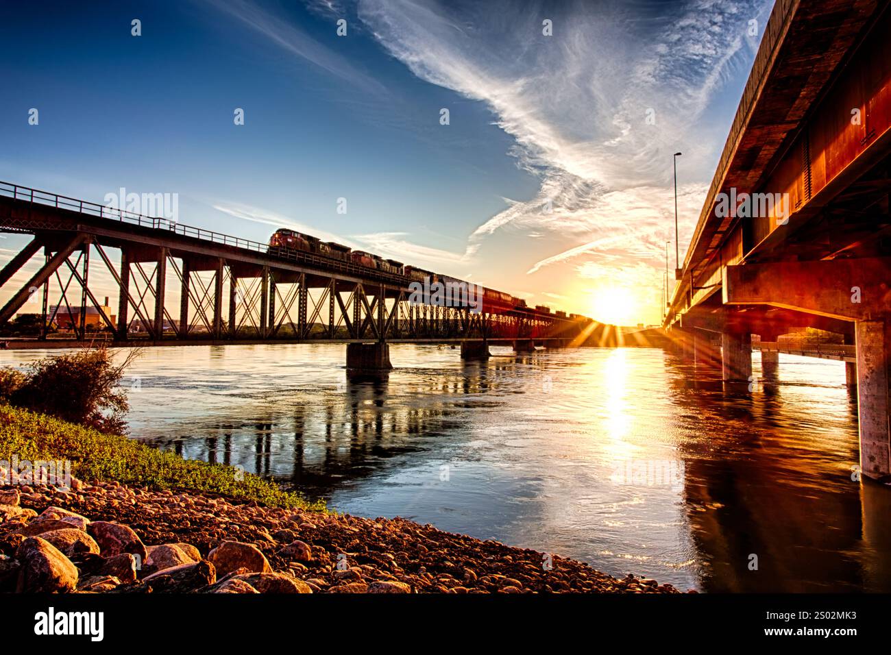 Un ponte attraversa un fiume con un treno che lo attraversa. Il sole sta tramontando, proiettando un caldo bagliore sulla scena Foto Stock