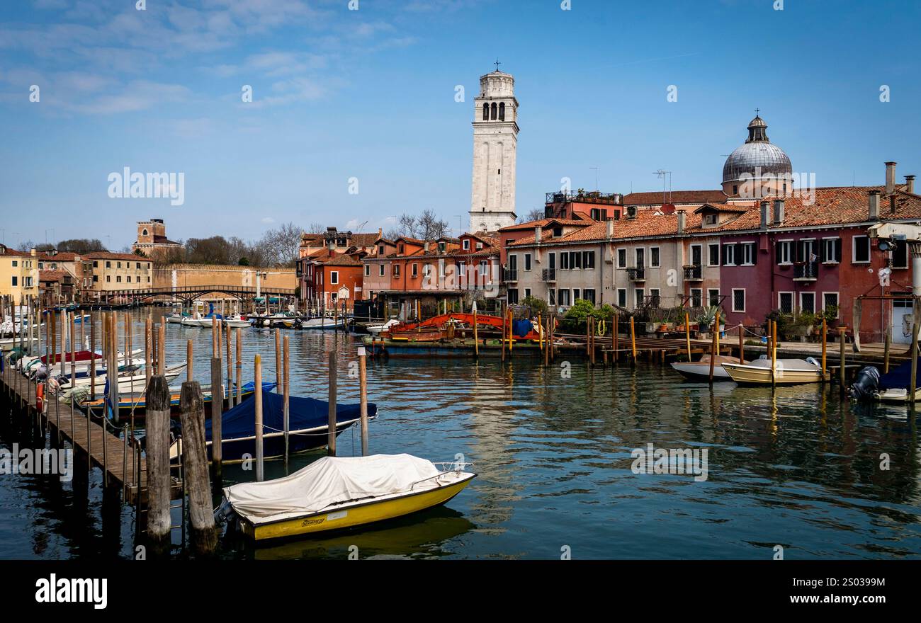 Una vista panoramica di un canale in una pittoresca cittadina italiana, caratterizzata da edifici colorati, barche ormeggiate lungo il molo e un alto campanile sullo sfondo Foto Stock