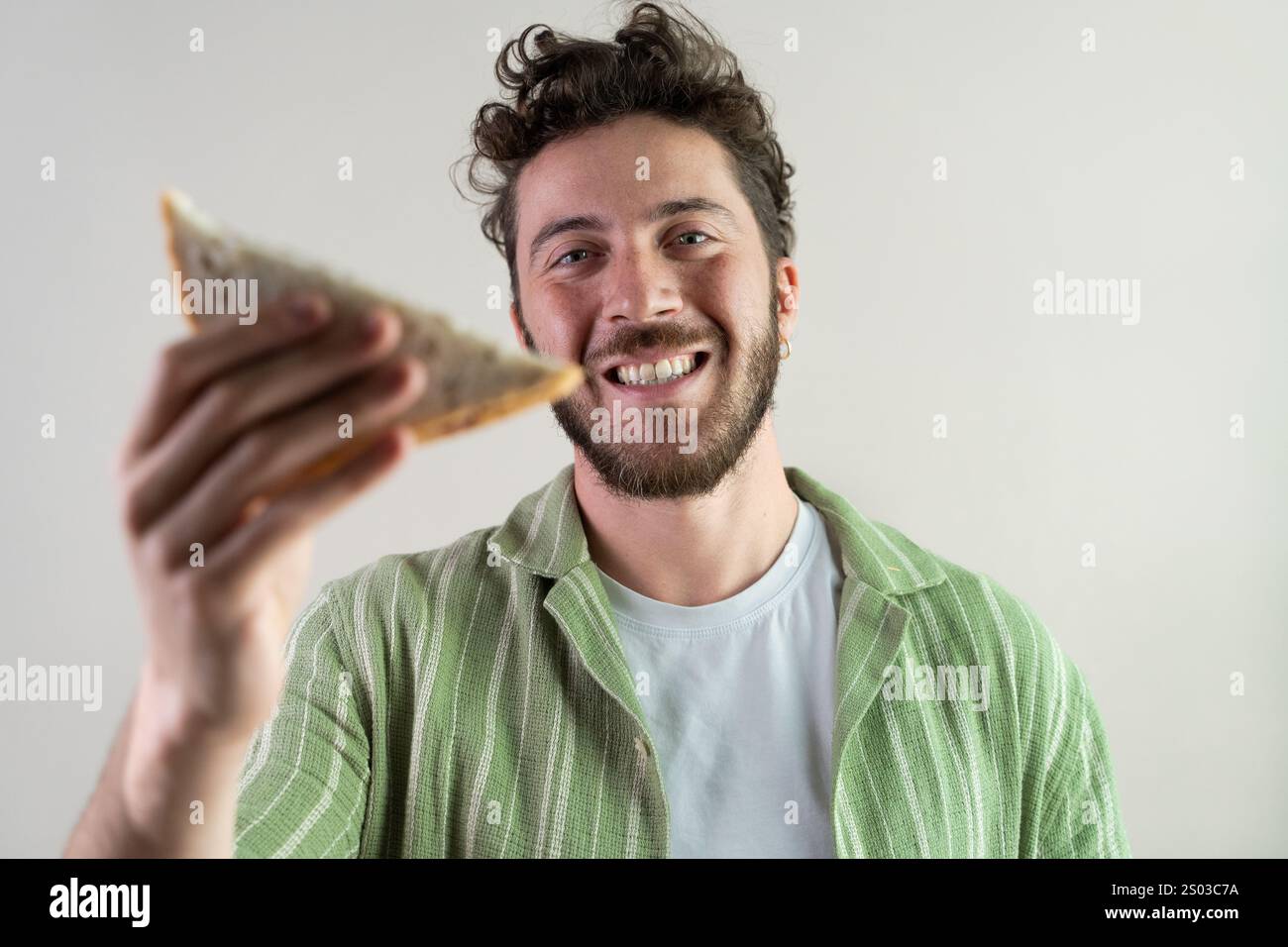 Un giovane sorridente che tiene un panino verso la macchina fotografica, godendosi un momento informale in casa. La scena enfatizza la felicità, il cibo e un'atmosfera rilassata. Foto Stock Un giovane sorridente che tiene un panino verso la macchina fotografica, godendosi un momento informale in casa. La scena enfatizza la felicità, il cibo e un'atmosfera rilassata. Foto Stock