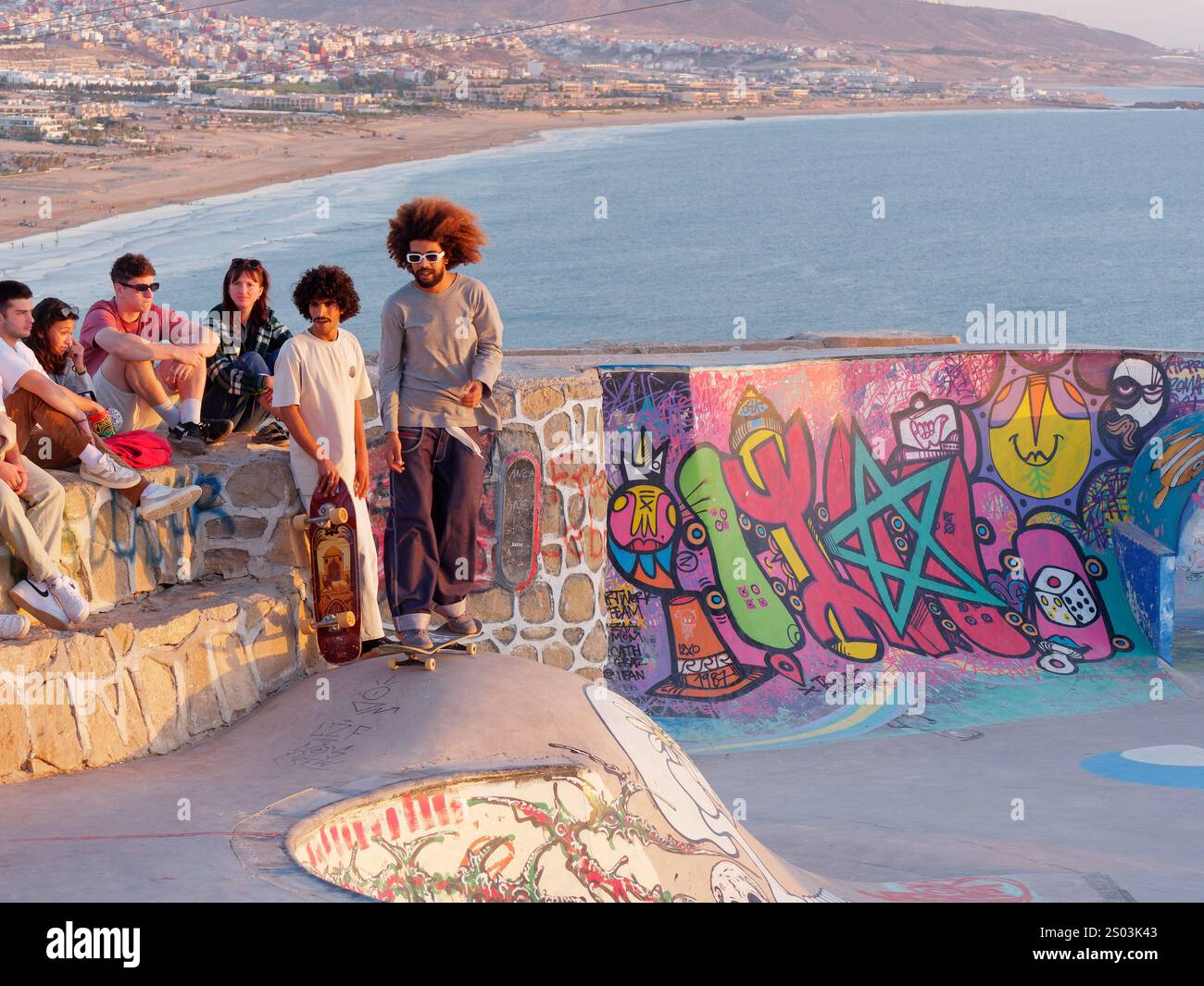 Due skateboard ti aspettano, girano accanto agli spettatori in uno Skate Park nel villaggio di Taghazout, con il mare e le montagne alle spalle, in Marocco. 23 dicembre 2024 Foto Stock