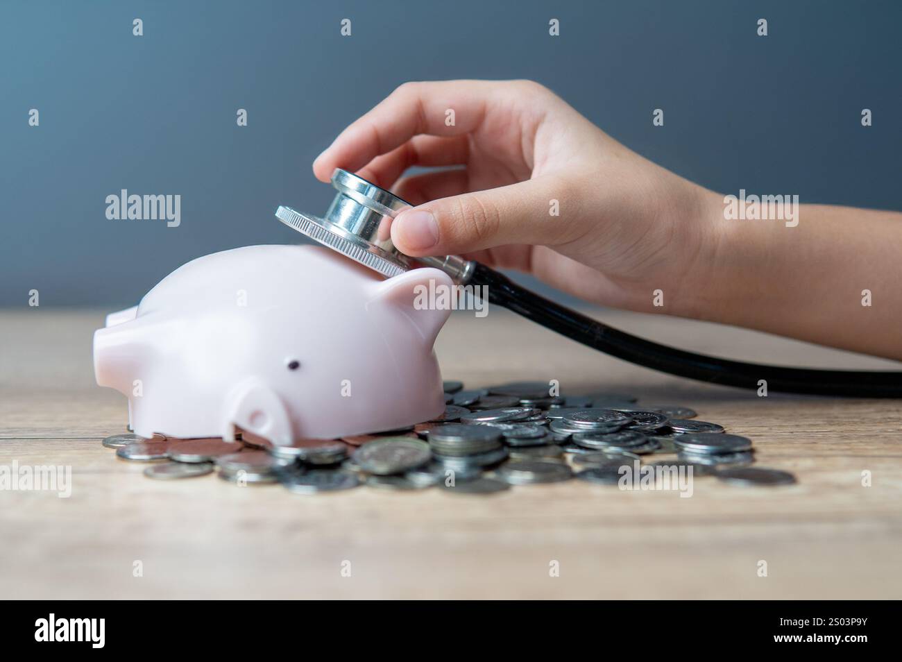 Una mano che regge uno stetoscopio che ascolta un salvadanaio rotto che giace su un mucchio di monete. Concetto di stato finanziario e di stato finanziario. Foto Stock