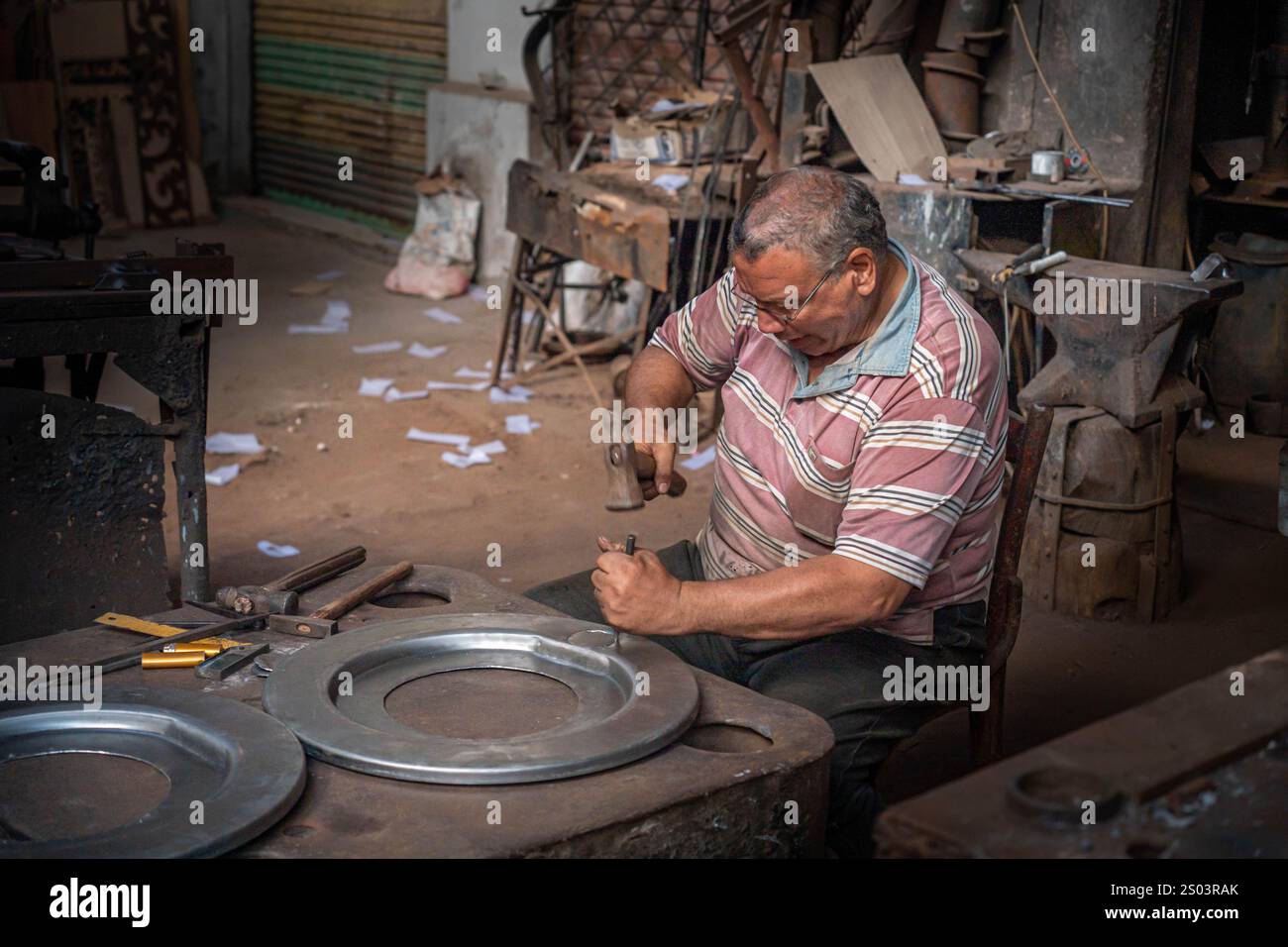 Un artigiano ad Alessandria, in Egitto, che lavora in un laboratorio, modellando meticolosamente una piastra di metallo a mano, circondata da strumenti e materiali tradizionali. Foto Stock