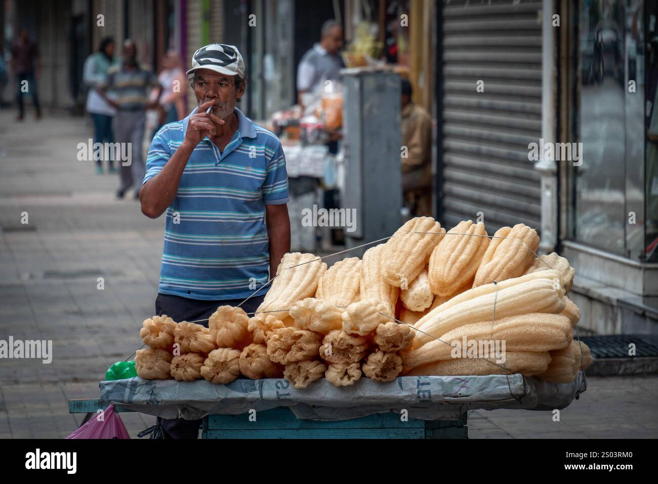 Venditore ambulante ad Alessandria, Egitto, fumare mentre vende loofah naturali. La scena cattura l'essenza della vita quotidiana nell'ambiente urbano. Foto Stock