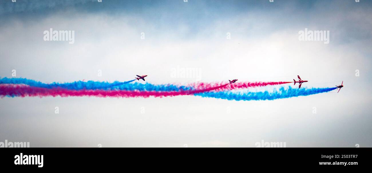 Mostra aerea con quattro aerei che eseguono una manovra sincronizzata, lasciando tracce di fumo rosso e blu contro un cielo nuvoloso. Foto Stock
