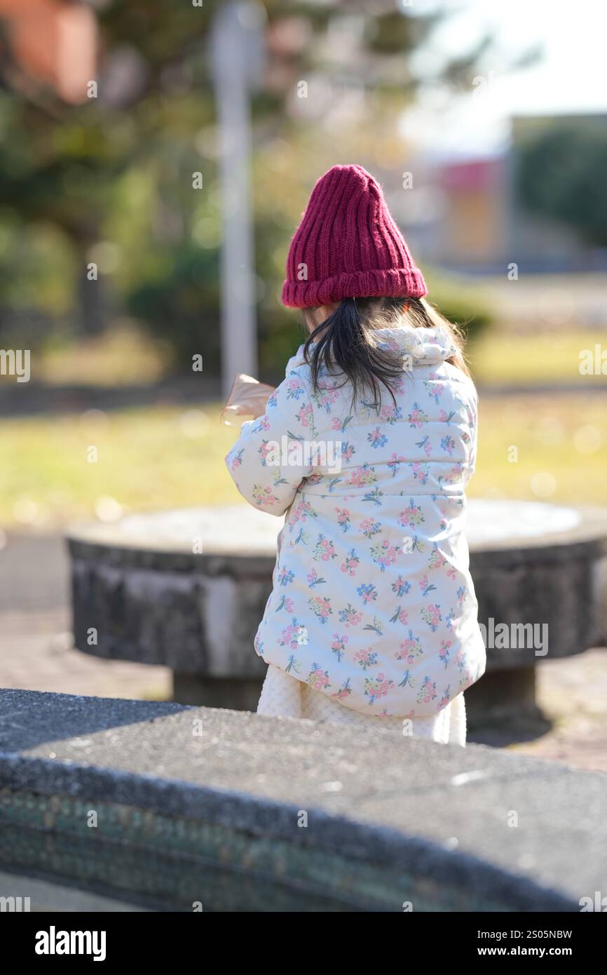 Una ragazza giapponese di 3 anni che indossa un cappello rosso a maglia e un piumino bianco cammina attraverso un parco di Hakata, nella prefettura di Fukuoka, in un freddo dopono invernale Foto Stock