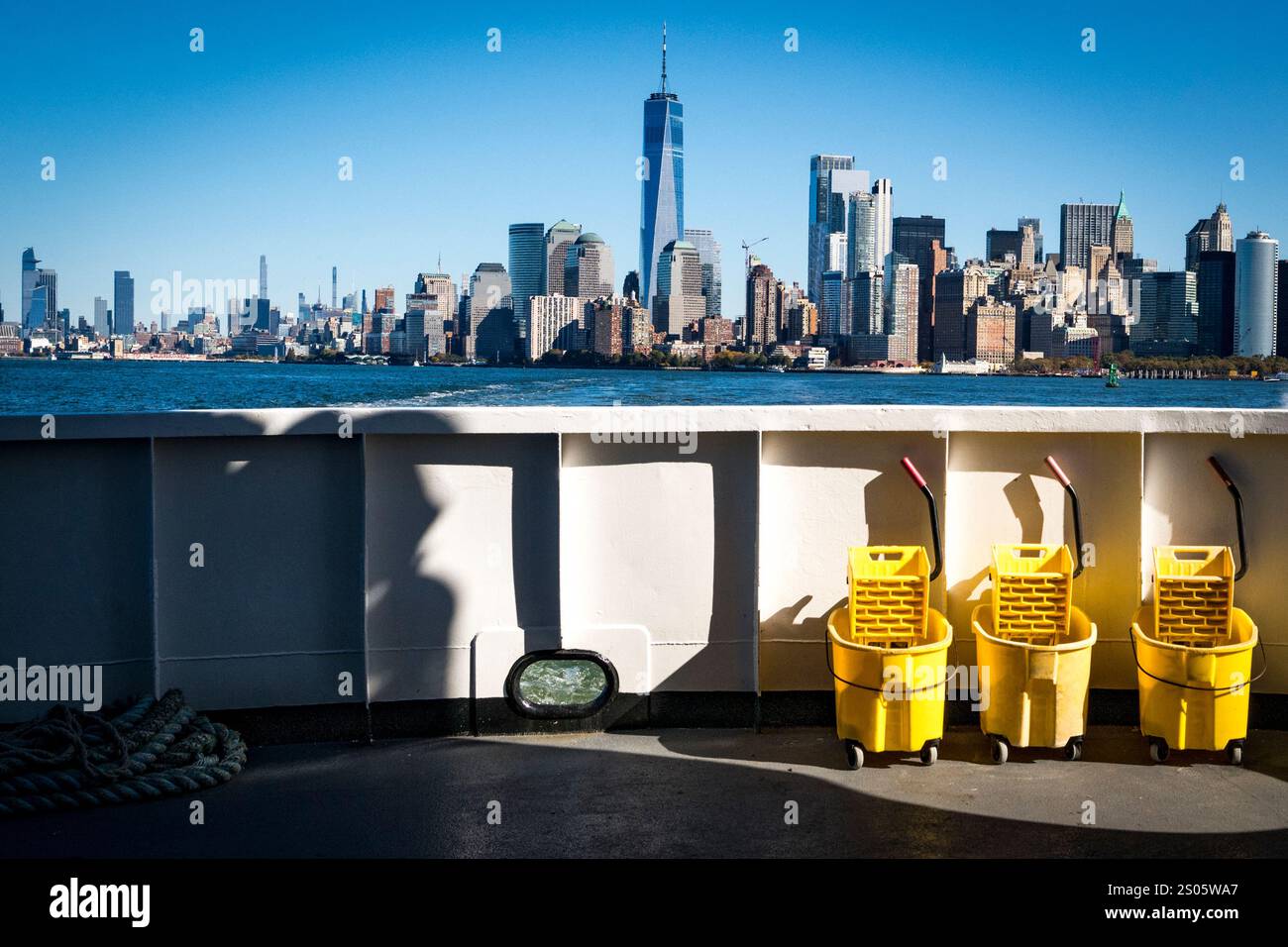 Prospettiva unica di Lower Manhattan e One World Trade Center, catturata dal traghetto di Liberty Island, incorniciata da carrelli gialli per le pulizie e un cielo blu. Foto Stock