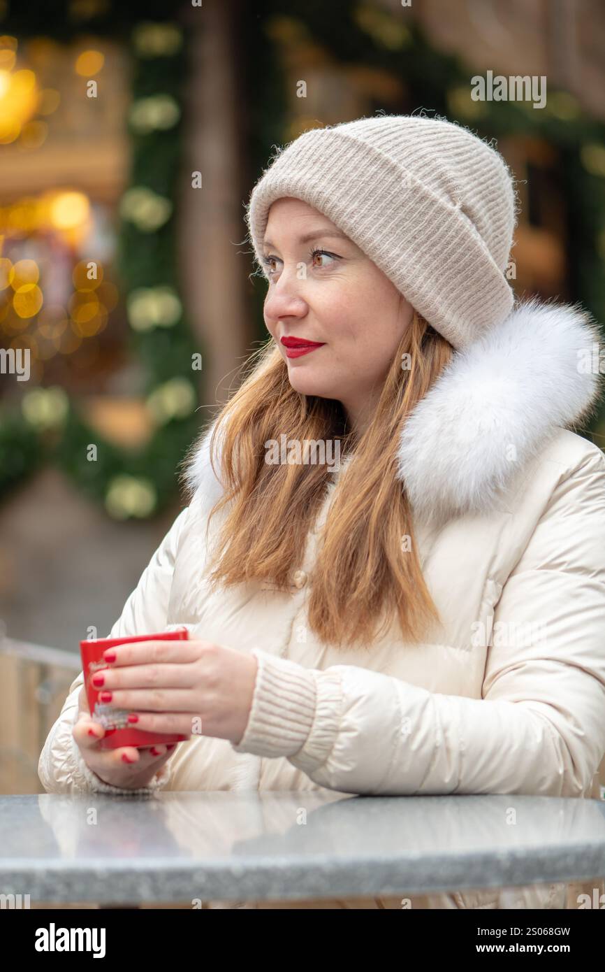 Una donna che regala una bevanda calda, vestita con un intimo cappotto invernale e perline, circondata da luci scintillanti e decorazioni festose in un'atmosfera urbana. Foto Stock