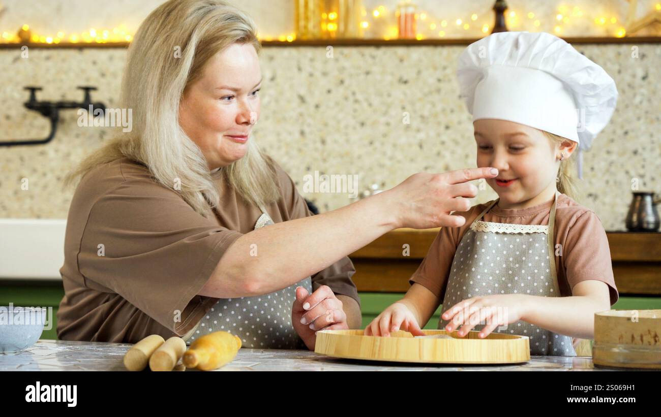 La madre sorridente spalma giocosamente la farina sul naso delle sue figlie creando un momento divertente e gioioso in cucina Foto Stock