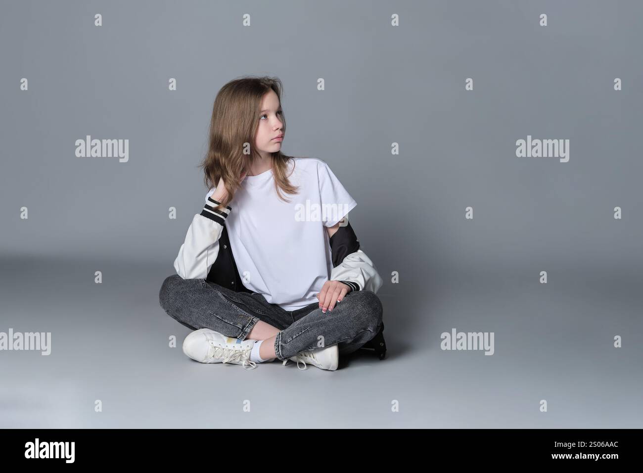 Una ragazza con un outfit casual si siede a gambe incrociate, perduta nel pensiero. Questa immagine mette in evidenza la miscela di innocenza e maturità emergente, simboleggiando il giurato Foto Stock