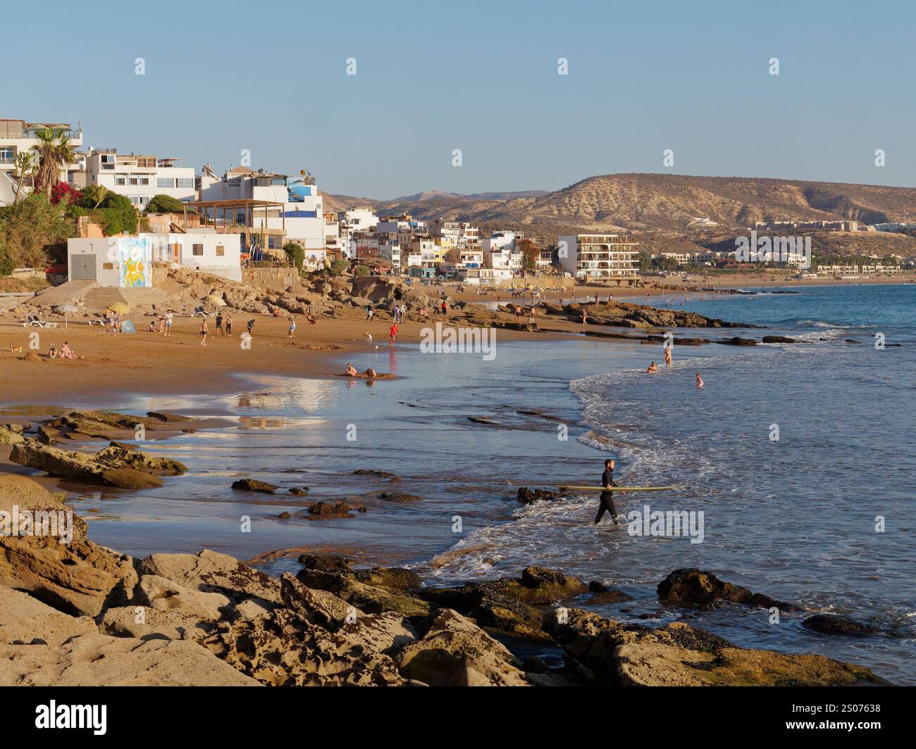 Turisti su una spiaggia rocciosa e sabbiosa mentre le onde si infrangono con il villaggio alle spalle di Taghazout, Marocco. 25 dicembre 2024. Foto Stock