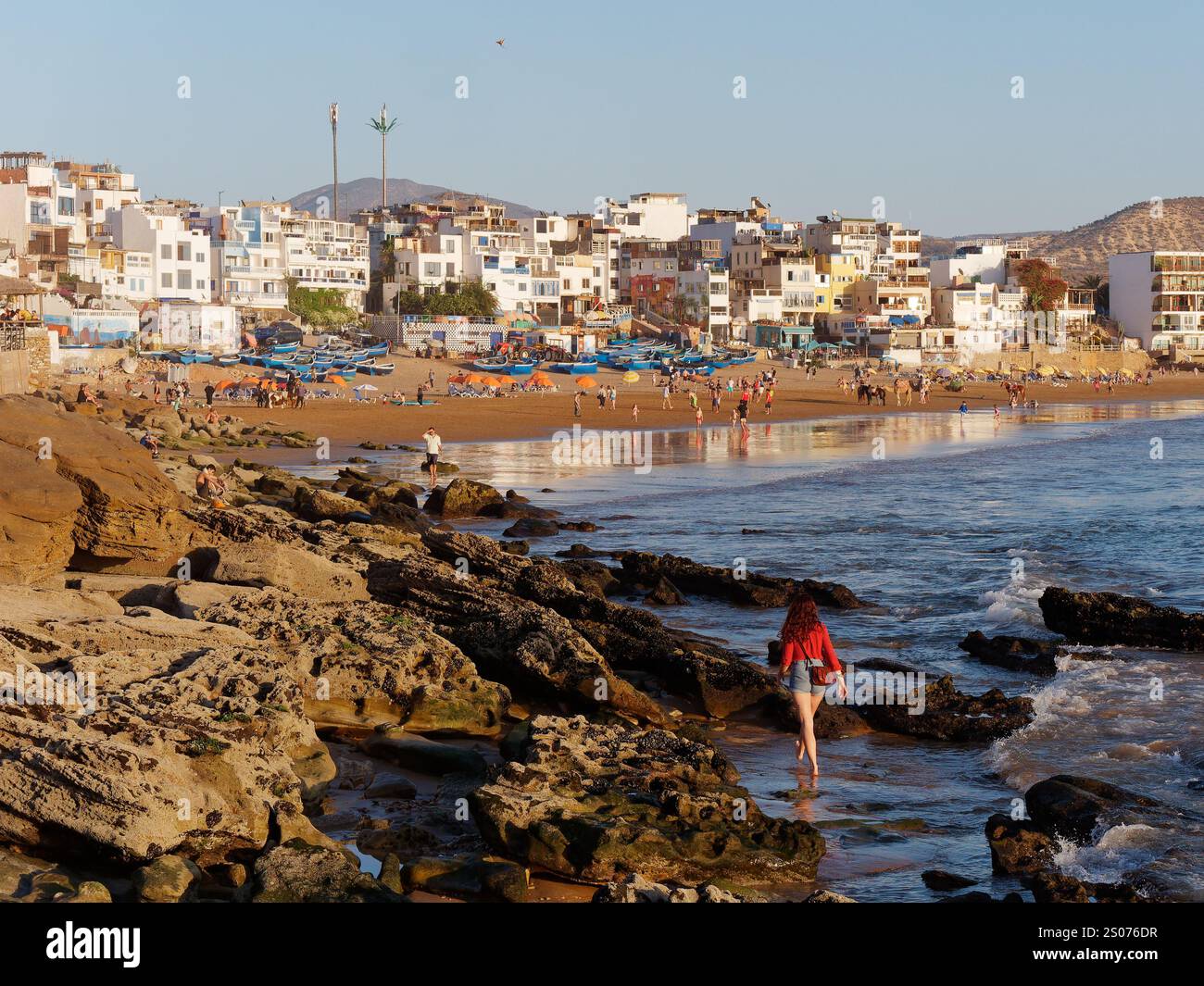 Spiaggia sabbiosa con barche da pesca blu come donna in passeggiate rosse lungo la costa rocciosa nel villaggio di pescatori di Taghazout, in Marocco. 25 dicembre 2024. Foto Stock