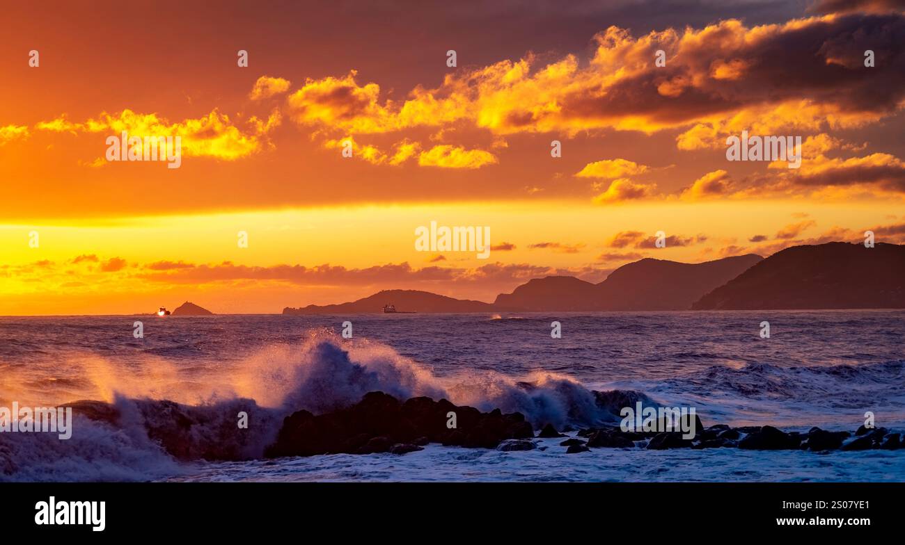 Uno splendido tramonto sull'oceano, con vivaci sfumature arancioni e gialle che illuminano il cielo. Le onde si infrangono contro le coste rocciose, mentre le isole lontane sono vicine Foto Stock