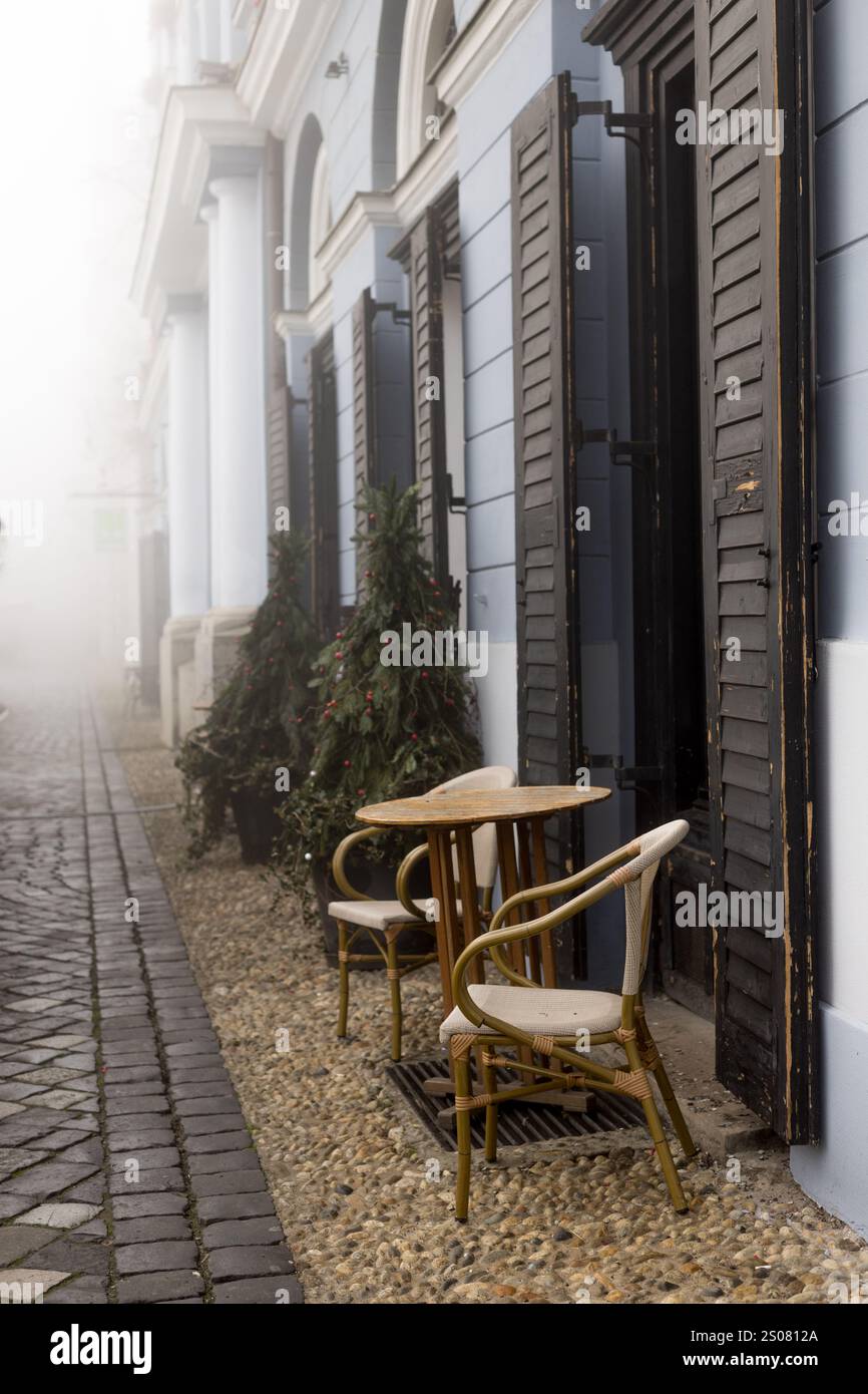 Una tranquilla scena mattutina con un tavolino da caffè in legno e sedie, sullo sfondo di una strada nebbiosa Foto Stock