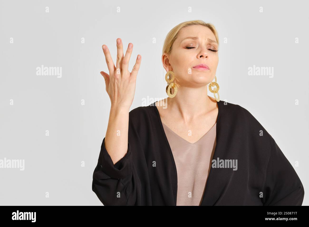 Una donna sta in piedi con gli occhi chiusi e la mano alzata in un gesto di frustrazione. Indossa orecchini eleganti e un outfit alla moda, chiaramente trasportabile Foto Stock