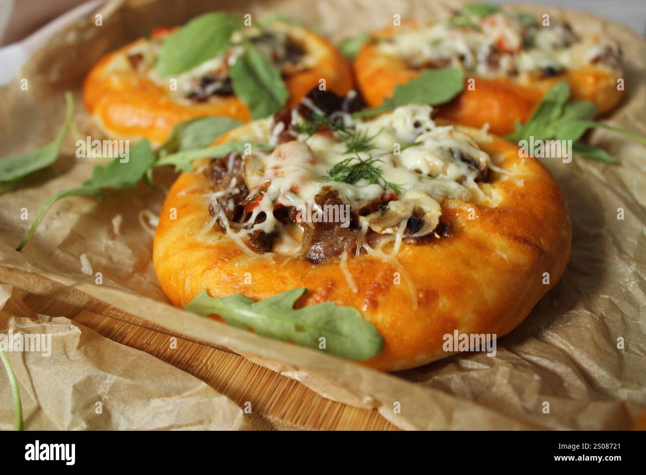 Mini pizze con funghi, manzo, cipolle, rucola e formaggio fuso poggiate su carta da forno Foto Stock