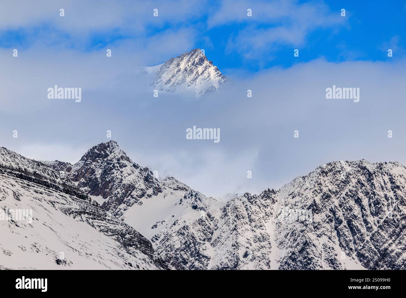 grazioso paesaggio di montagne rocciose coperte di neve sormontate da nuvole, ma sopra le nuvole si trova una vetta più alta in una zona di cielo blu nella georgia meridionale Foto Stock