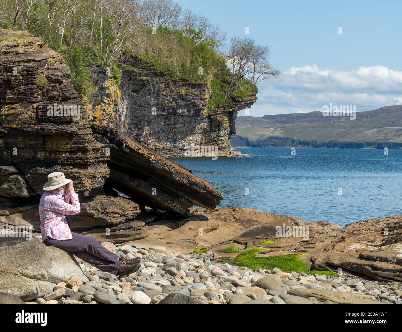 Donna adulta che guarda la fauna selvatica attraverso un binocolo su una spiaggia rocciosa in una giornata di sole a maggio, Glasnakille vicino a Elgol, Isola di Skye, Scozia, Regno Unito Foto Stock