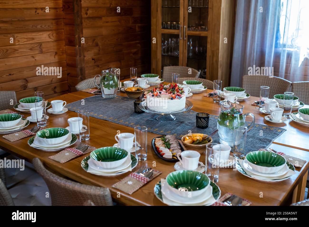 Un intimo interno in legno presenta un tavolo da pranzo con una torta alla frutta, stoviglie verdi e bianche e antipasti. La luce naturale filtra attraverso il trasparente Foto Stock