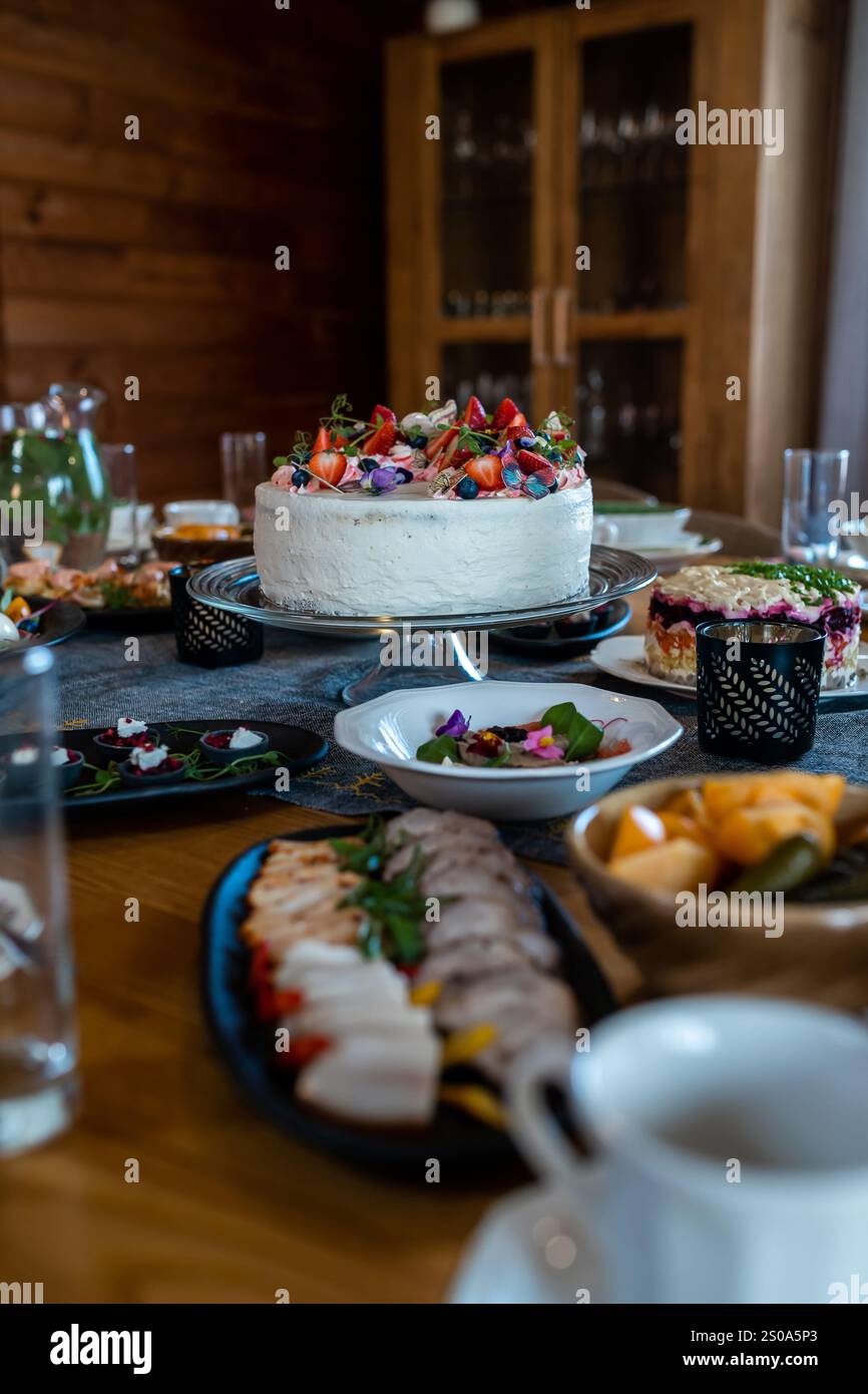 Un tavolo da pranzo interno e' allestito con una grande torta ricoperta di frutti di bosco e fiori, circondato da antipasti e contorni, con un armadietto di legno vicino Foto Stock