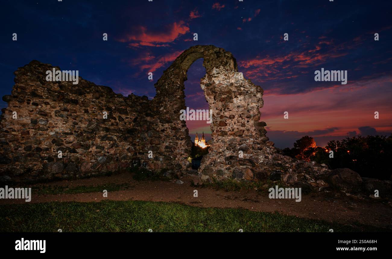 Sagoma di un'antica arcata in pietra contro un vivace cielo serale a Rezekne, Lettonia. Un edificio illuminato con guglie è visibile attraverso l'arco. Foto Stock