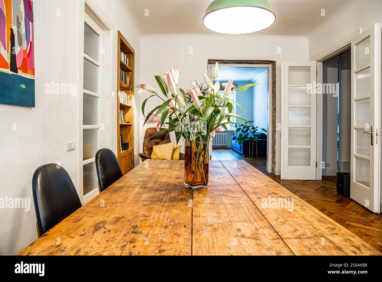 Un rustico tavolo da pranzo in legno con fiori rosa e' illuminato da una lampada a soffitto. L'arte moderna decora le pareti e una libreria aggiunge calore allo spazio. Foto Stock