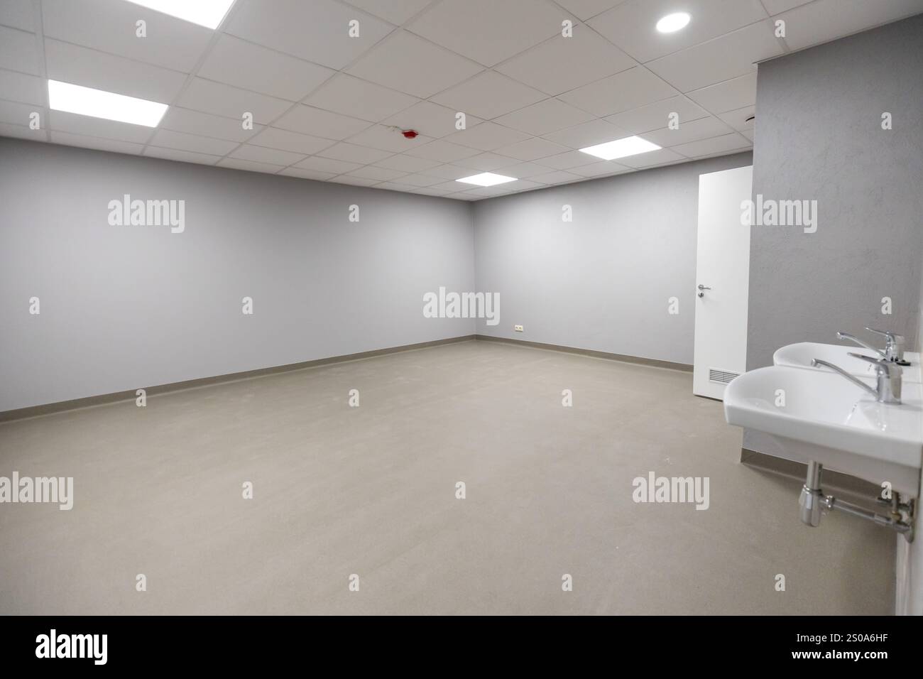 Una camera vuota presenta pareti grigie neutre, pavimento di colore chiaro, illuminazione a incasso, porta bianca e doppio lavabo montato a parete. Foto Stock