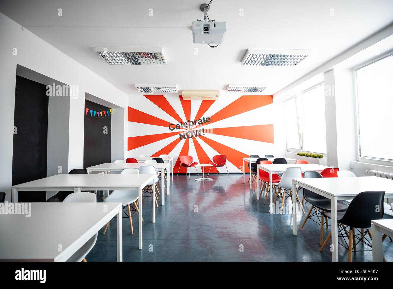 Una sala luminosa presenta un suggestivo design a parete con raggi rossi e bianchi con "Celebrate NOW". Tavoli bianchi, sedie rosse e grandi finestre completano il Foto Stock