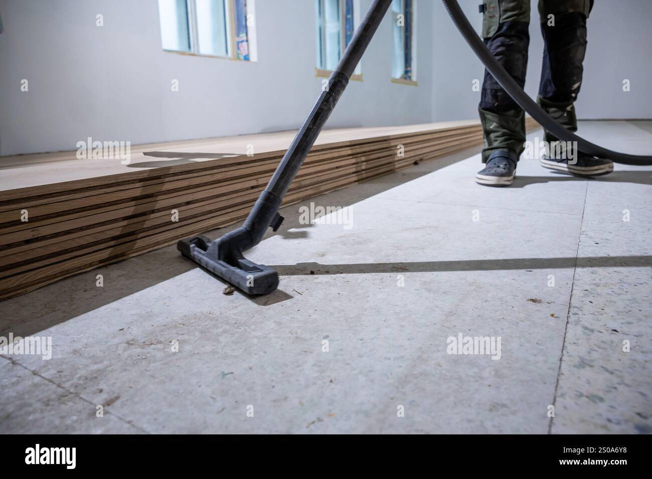 Un lavoratore pulisce il sottopavimento con un aspirapolvere in una stanza ben illuminata. I pavimenti in legno impilati in modo ordinato sono visibili, con luce naturale che scorre attraverso WIN Foto Stock