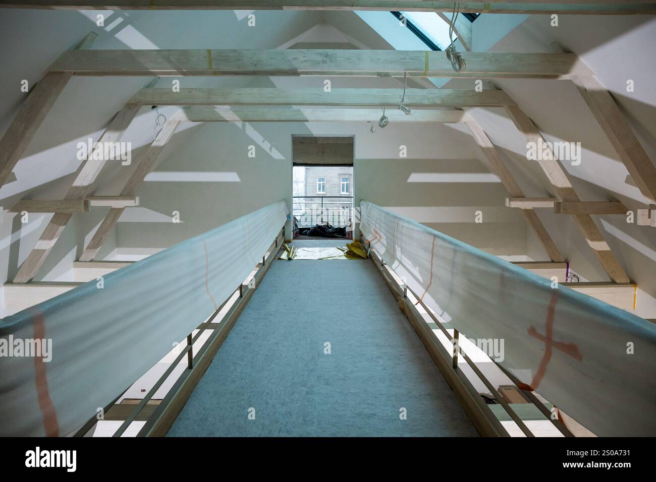 Una stretta passerella in un edificio in costruzione presenta travi di legno esposte e un lucernario. Una finestra alla fine rivela un edificio urbano adiacente Foto Stock