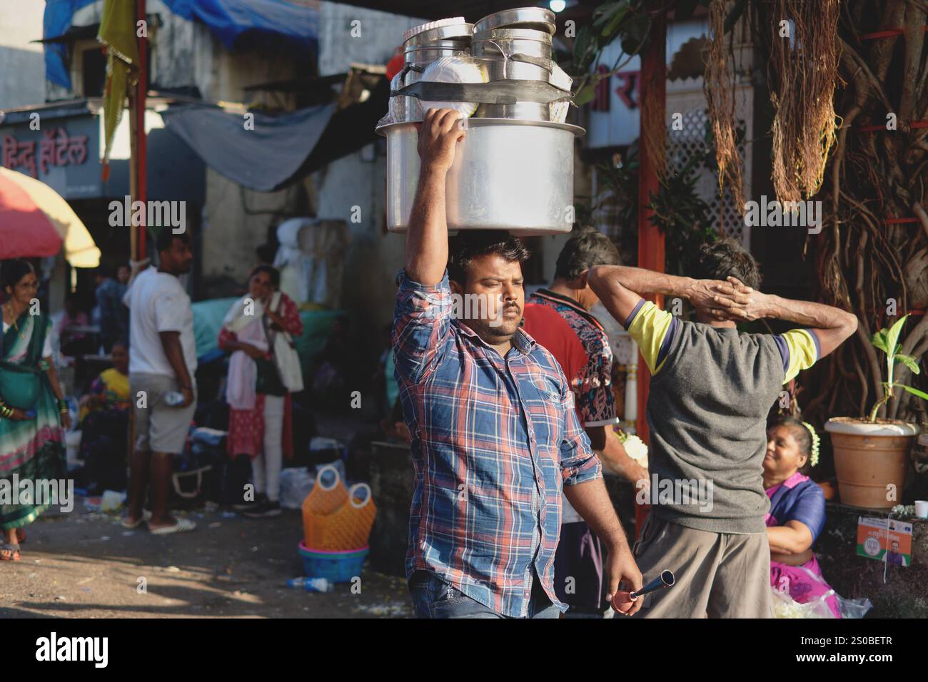 Un venditore ambulante di snack o cibo di strada a Mumbai, in India, che porta utensili, vasi e prodotti alimentari sulla testa, passeggiando attraverso un'affollata area del mercato Foto Stock