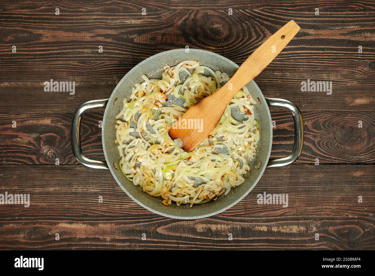 Le cipolle tritate e l'aglio vengono saltati in una padella con una spatola di legno, creando una base saporita per un pasto su un rustico tavolo di legno. La ripresa della scena Foto Stock