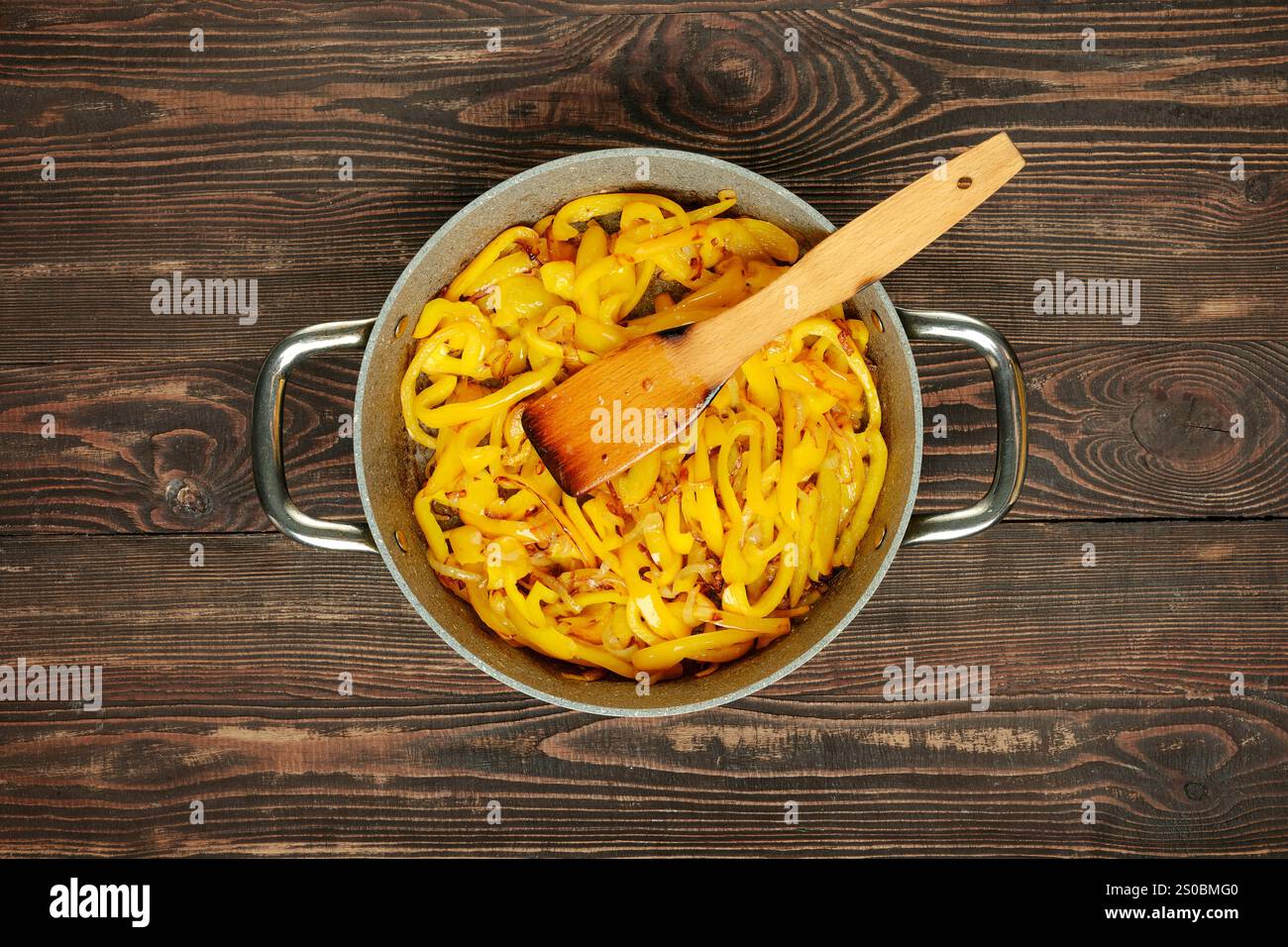 I peperoni gialli freschi vengono saltati in una padella con una spatola di legno, che mostra colori vivaci e tecniche di cottura. Il piano di appoggio in legno aggiunge un Foto Stock