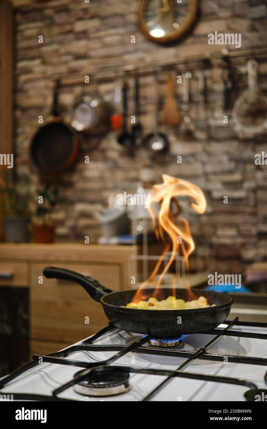 Le fiamme ballano da una padella sul fornello mentre le verdure vengono saltate in un'accogliente cucina. Il decor rustico presenta pareti in pietra e cucina ut Foto Stock
