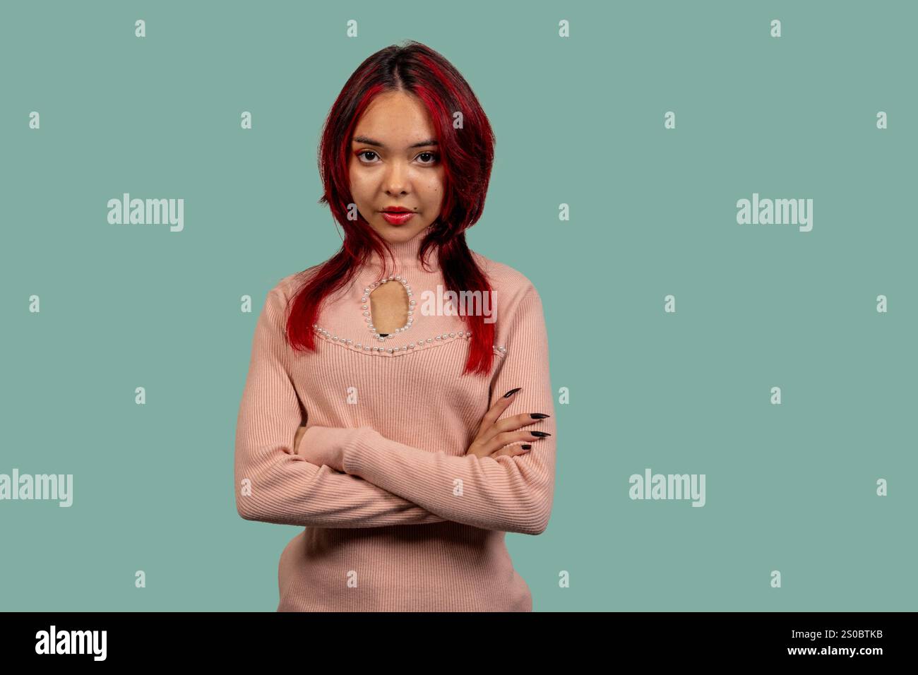 Ritratto in studio di una giovane donna sicura di sé con i capelli rossi tinti, che indossa un elegante pullover, con le braccia incrociate su un vivace dorso turchese Foto Stock