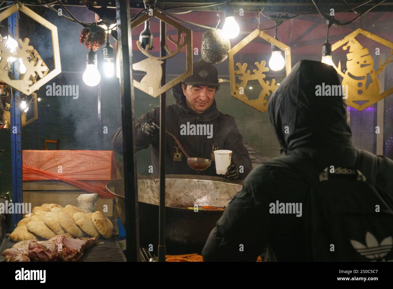 Un venditore serve vino caldo fumante in un mercatino di Natale vicino a NDK a Sofia, Bulgaria. La scena si illumina di luci e decorazioni natalizie. Foto Stock
