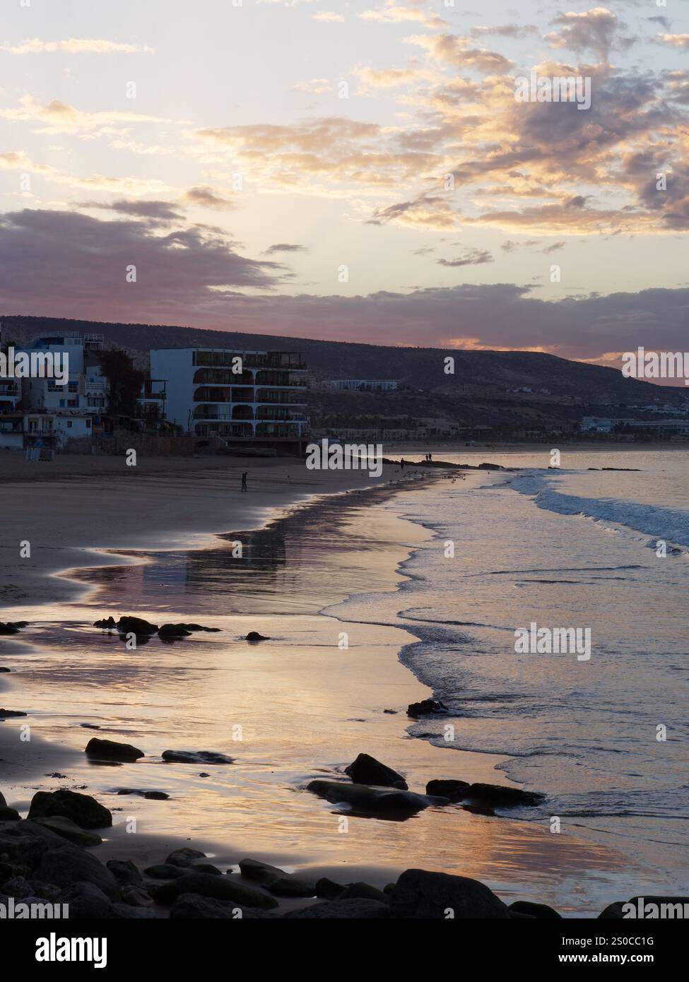 Cielo rosso all'alba sopra il villaggio di Taghazout e con la sua spiaggia sabbiosa e rocciosa mentre le dolci onde si infrangono in Marocco. 27 dicembre 2024. Foto Stock