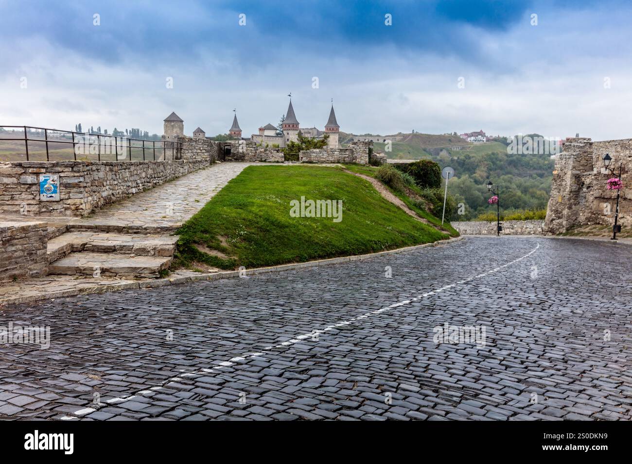 Una strada in pietra con un castello sullo sfondo. Il castello è circondato da un muro e ha una torre. La strada è bagnata e ha una superficie acciottolata Foto Stock Una strada in pietra con un castello sullo sfondo. Il castello è circondato da un muro e ha una torre. La strada è bagnata e ha una superficie acciottolata Foto Stock