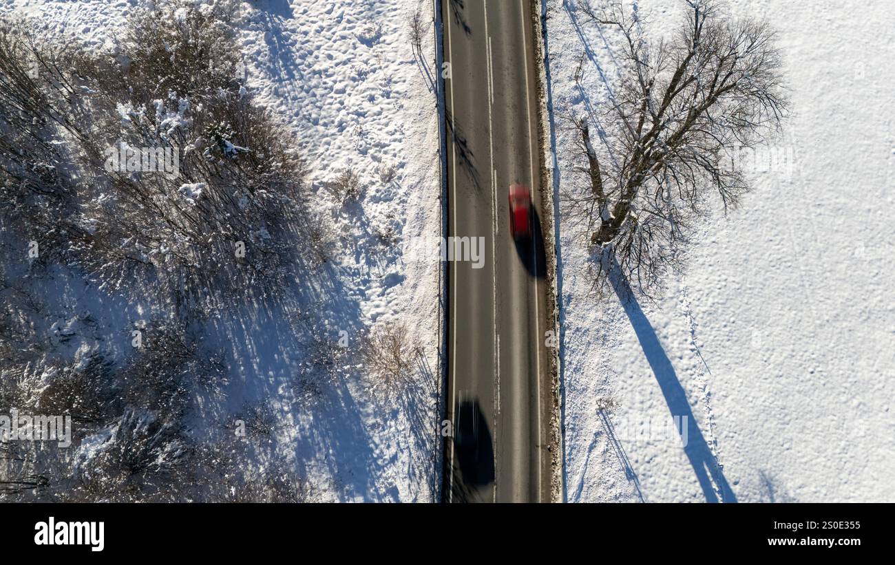 Una vista aerea dinamica che cattura un'auto rossa in movimento su una strada rurale fiancheggiata da campi innevati e alberi. Foto Stock
