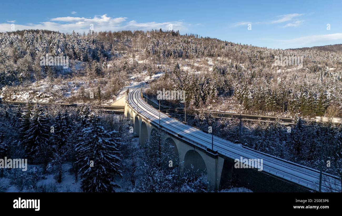 Una vista aerea panoramica di un ponte ferroviario coperto di neve che si snoda attraverso una fitta foresta invernale, con cieli azzurri e un tranquillo sfondo montano. Foto Stock