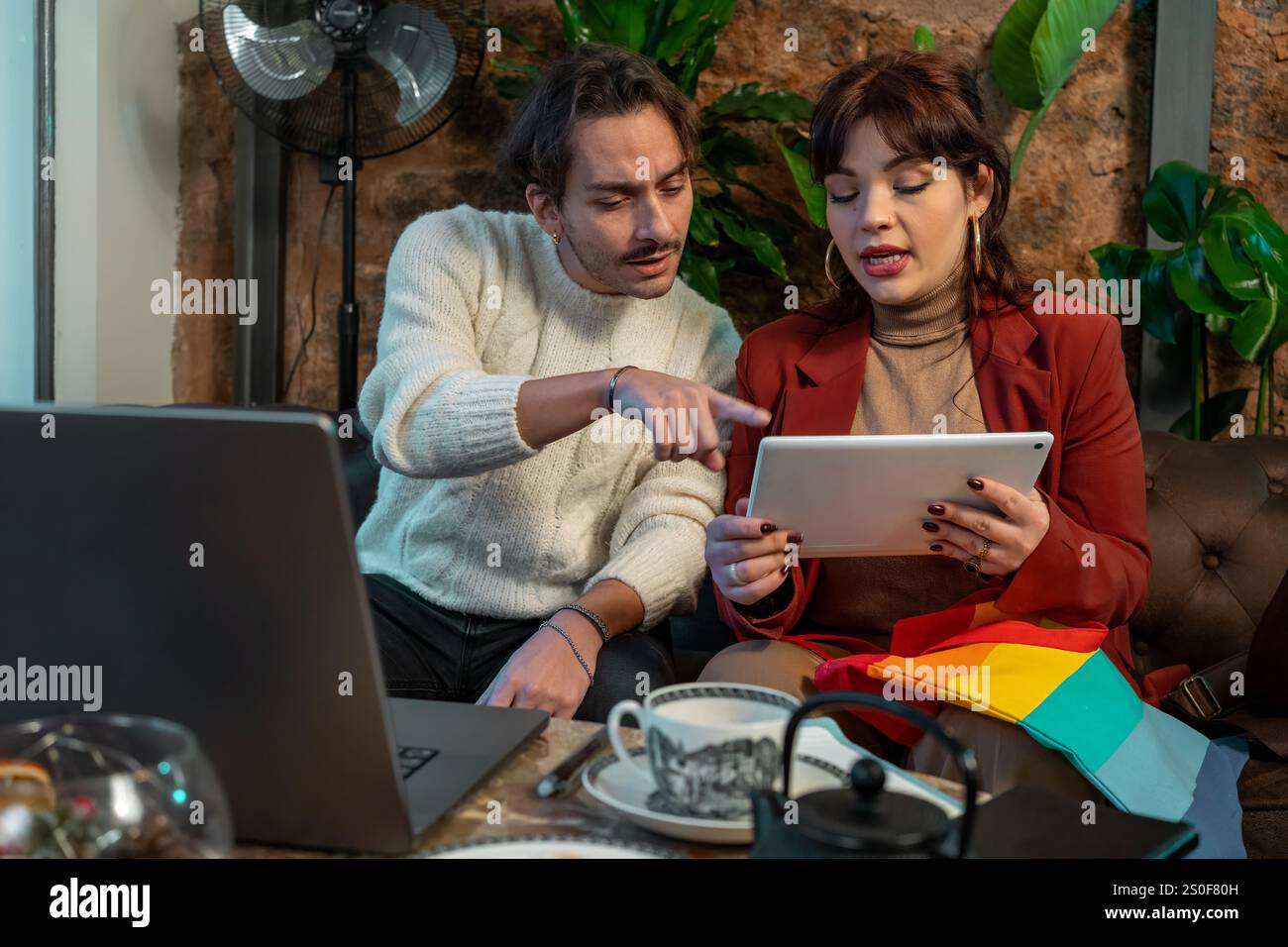 Un uomo e una donna che lavorano insieme con un tablet e un notebook in un accogliente tavolino circondato da piante, concentrandosi sulla collaborazione e sul lavoro di squadra. Foto Stock