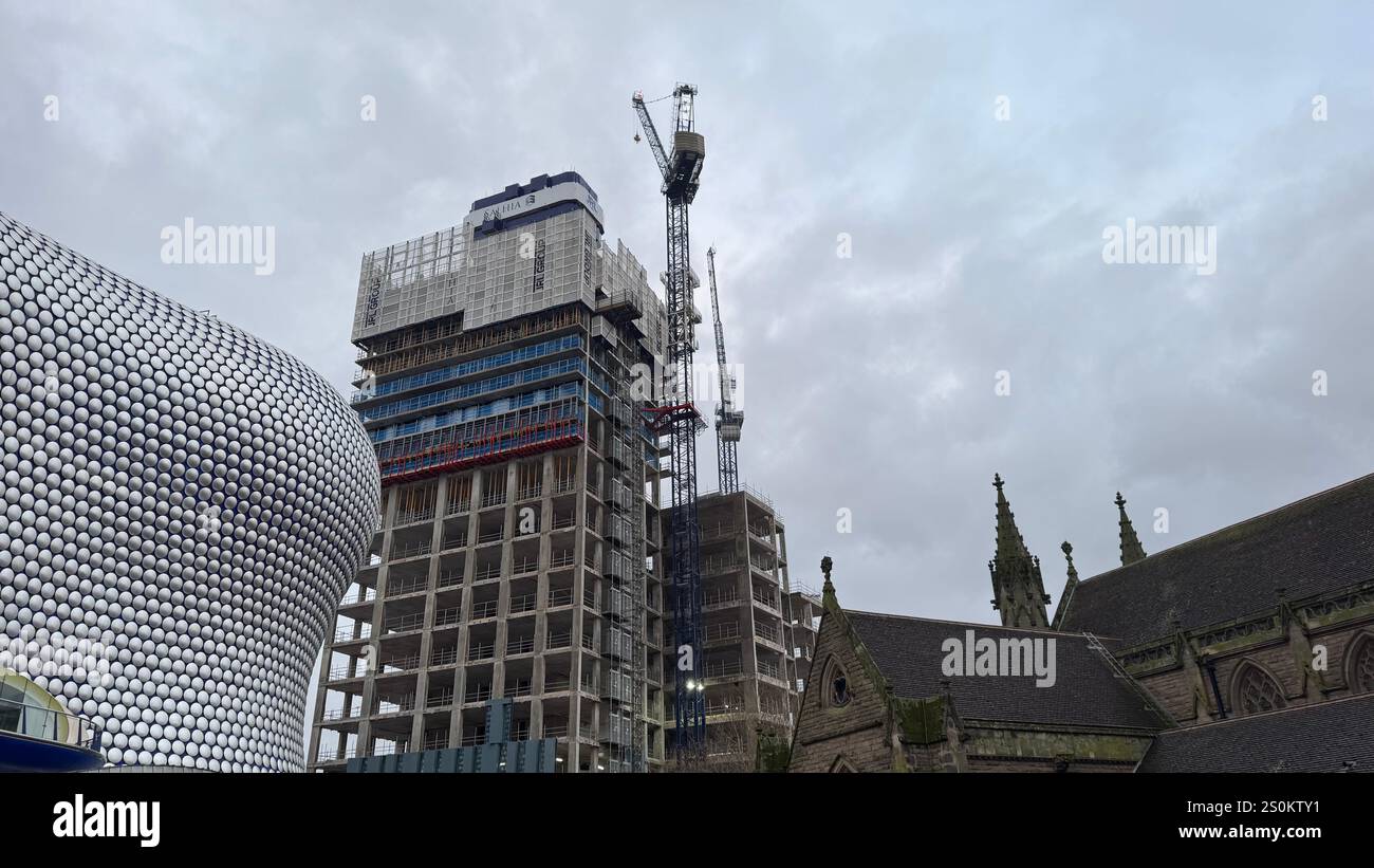 Sotto un cielo nuvoloso, un alto edificio parzialmente costruito sorge accanto all'edificio Selfridges e una parte di una chiesa. Gru da cantiere Foto Stock