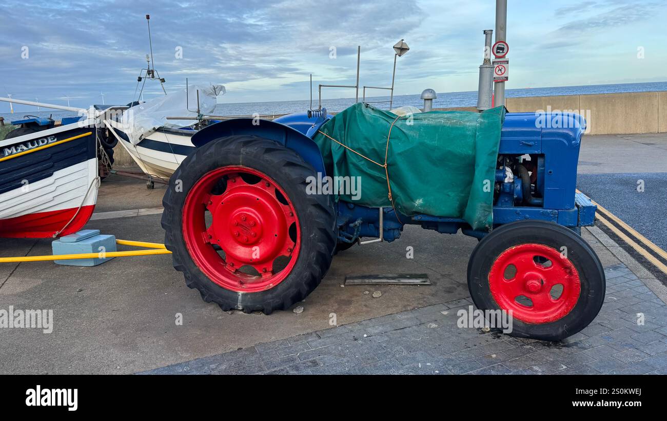 Un trattore blu con ruote rosse, parzialmente coperto da un telone verde, è parcheggiato vicino a una piccola barca da pesca chiamata "Maude". La scena è ambientata all'aperto da l Foto Stock