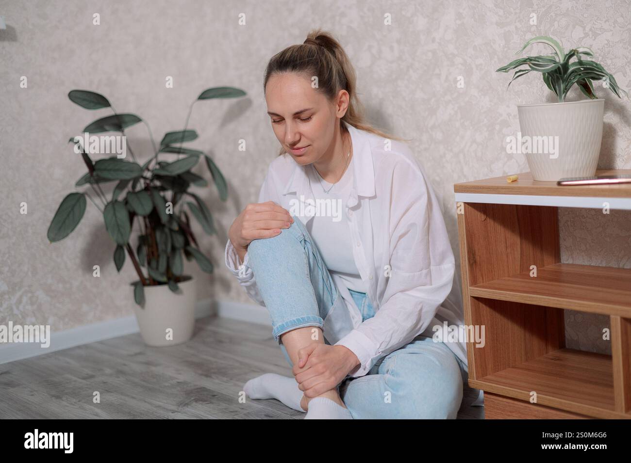 Una giovane donna vestita in modo informale è seduta su un divano grigio, sembra sentirsi disagio nel ginocchio. Regola la sua postura con cura in modo sereno, Foto Stock